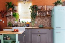 a bright kitchen with orange walls, a bright printed tile backsplash, a blue fridge, a green kitchen island and potted greenery