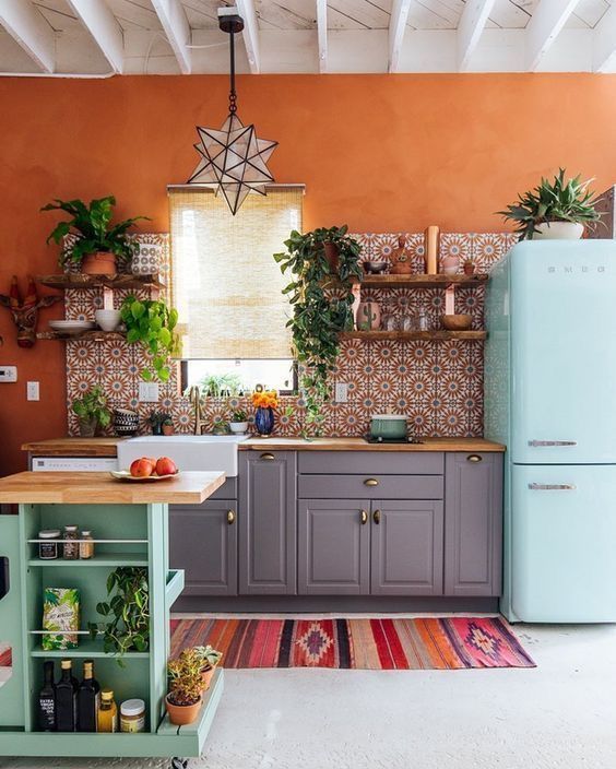 a bright kitchen with orange walls, a bright printed tile backsplash, a blue fridge, a green kitchen island and potted greenery