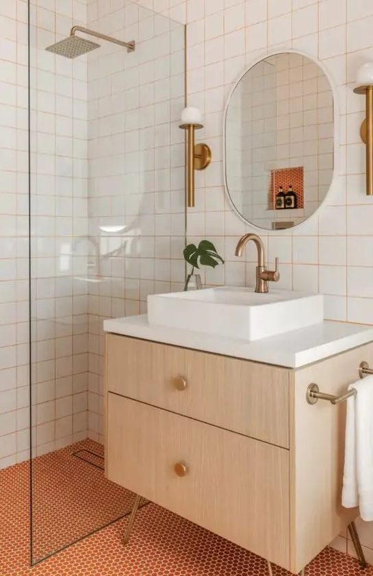 a bright modern bathroom with white square tiles with orange grout and orange penny tiles, a timber vanity, an oval mirror and brass fixtures