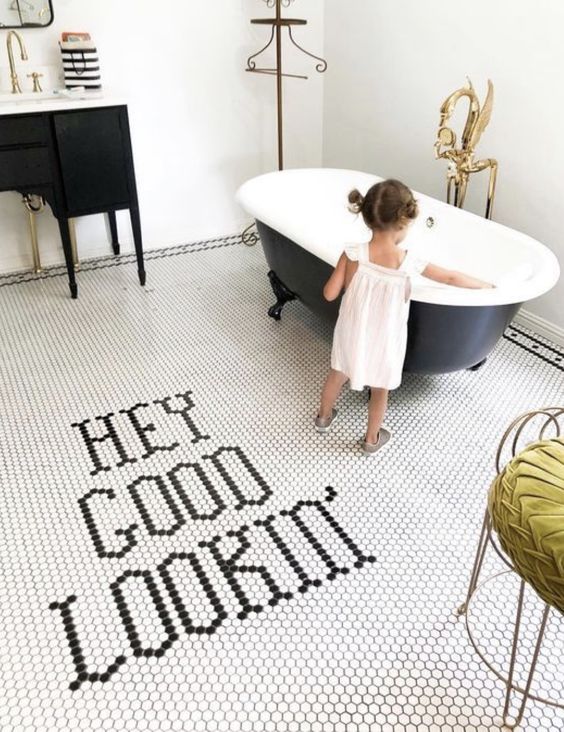 a chic bathroom with white walls, a white penny tile floor with letters, a black tub and a black vanity, gold touches