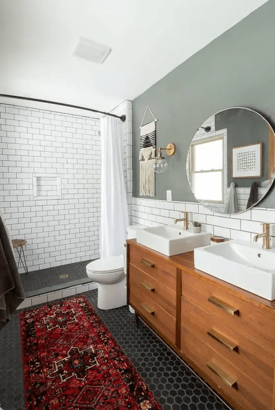 a chic mid-century modern bathroom with a grey accent wall penny and subway tiles, a stained vanity, a round mirror and two sinks
