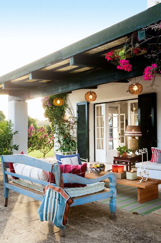 a colorful Mediterranean terrace with bright blooms, pendant lamps, a blue bench and colorful textiles plus greenery