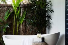 a contemporary luxe outdoor bathroom with shutters over it and a natural wall, greenery and a chic oval tub