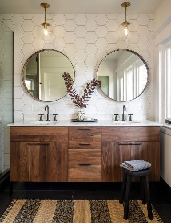 A cool mid century modern bathroom with white hex tiles, a stained vanity, round mirrors, pendant lamps, a striped rug