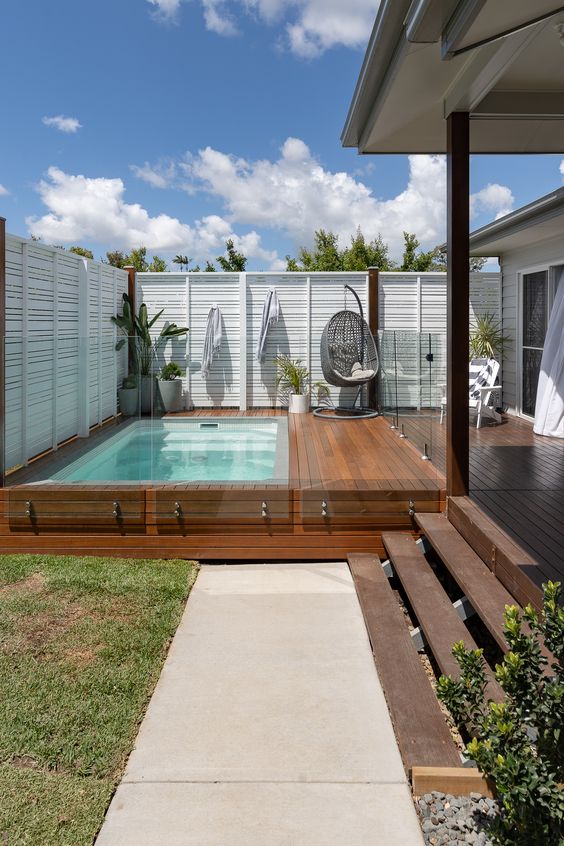 a cool outdoor modern space wiht a wooden deck and a plunge pool, some furniture and potted greenery and a glass fence around
