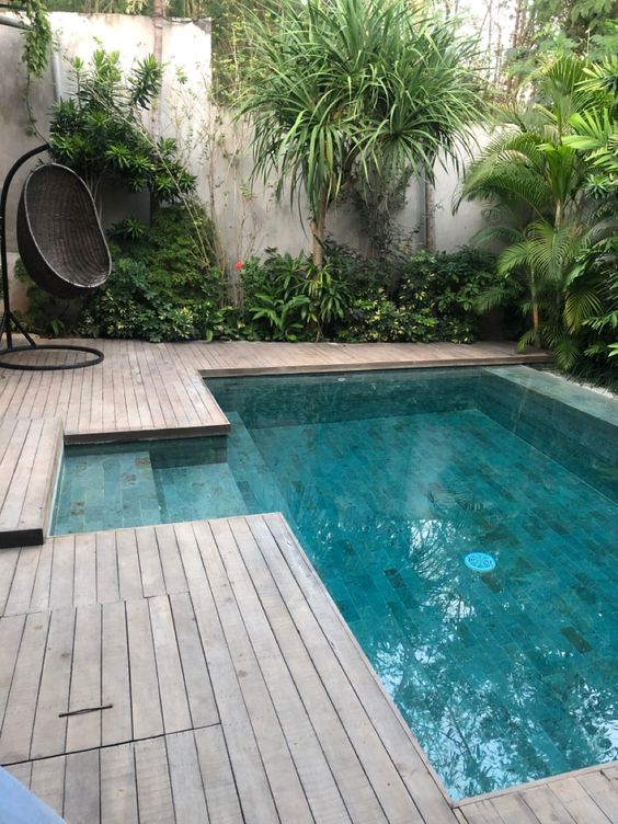 A cool tropical outdoor space with a wooden deck, trees and greenery around, a black woven egg shaped chair and a plunge pool is cool