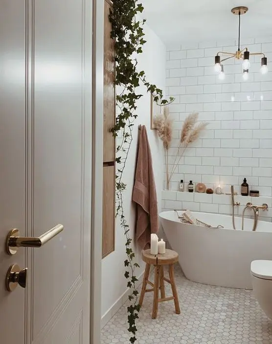 a cozy boho bathroom clad with white subway and marble hex tiles, a free-standing tub, potted greenery, pampas grass and a chandelier