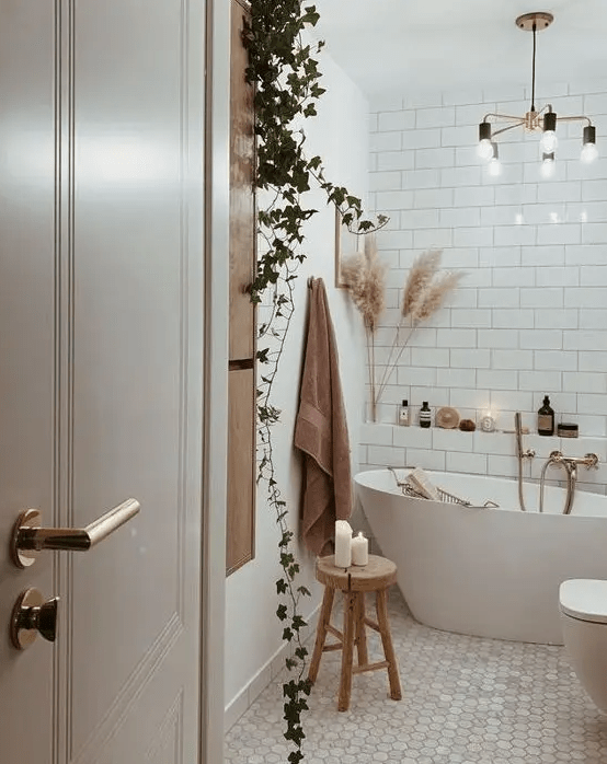 A cozy boho bathroom clad with white subway and marble hex tiles, a free standing tub, potted greenery, pampas grass and a chandelier