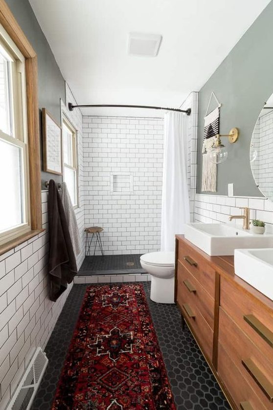 A cozy mid century modern bathroom with white subway and black hex tiles, with a wooden vanity, a boho rug and windows