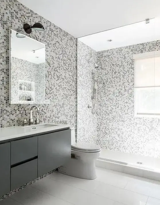 a grey and white bathroom with a window in the shower, grey and white penny tiles, a grey vanity, a mirror and white appliances