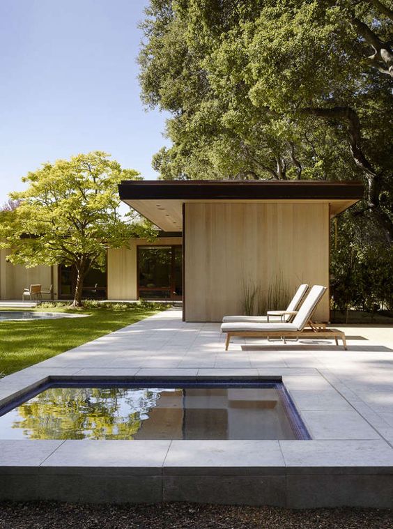 a minimalist outdoor space with a stone deck, a plunge pool, a couple of loungers and a green lawn around