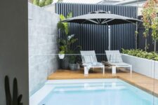 a modern backyard with a wooden deck, a plunge pool, some greenery and potted plants, loungers and an umbrella