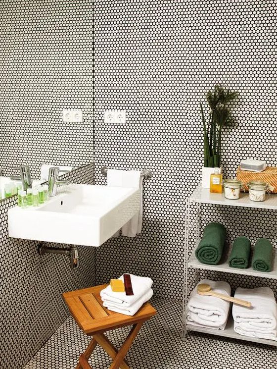 a modern bathroom clad with white penny tiles with black grout, a storage shelf with towels, a sink, a stool and some greenery