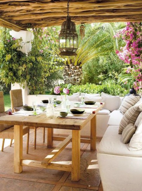a neutral Meidterranean terrace with a white L-shaped sofa, wooden furniture, Moroccan lamps and bright blooms