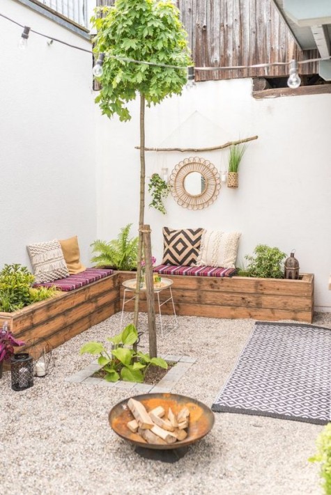 a neutral boho terrace with a large L-shaped wooden bench with greenery planted, a fire bowl and candle lanterns