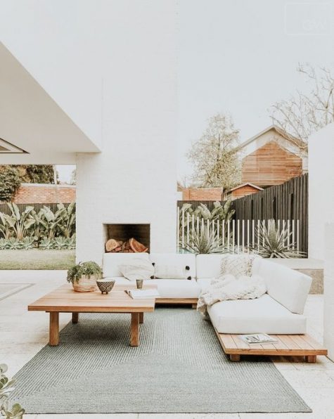 A neutral modern terrace with a fireplace, a pallet sofa and a table plus some greenery