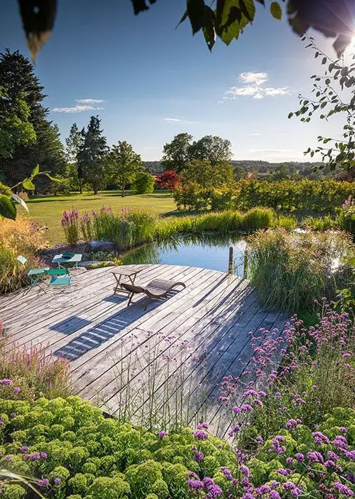 a pretty small swimming pond with water plants, a large wooden deck with loungers, chairs and a table and blooms and greenery around