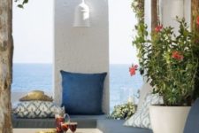 a sea Mediterranean terrace done in blue and white, with potted greenery and blooms, a concrete bench and printed pillows