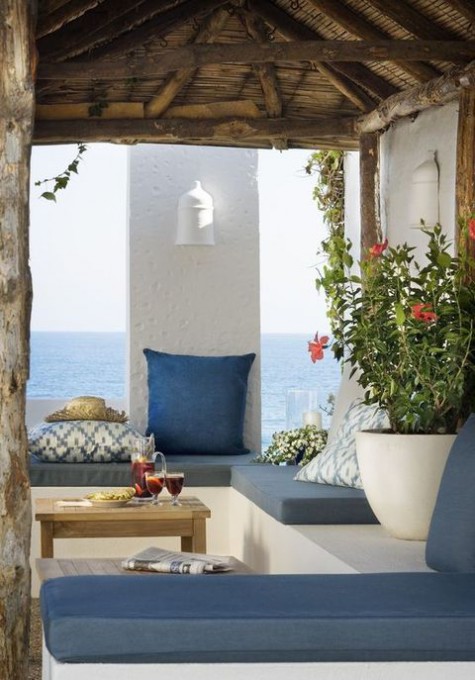 a sea Mediterranean terrace done in blue and white, with potted greenery and blooms, a concrete bench and printed pillows
