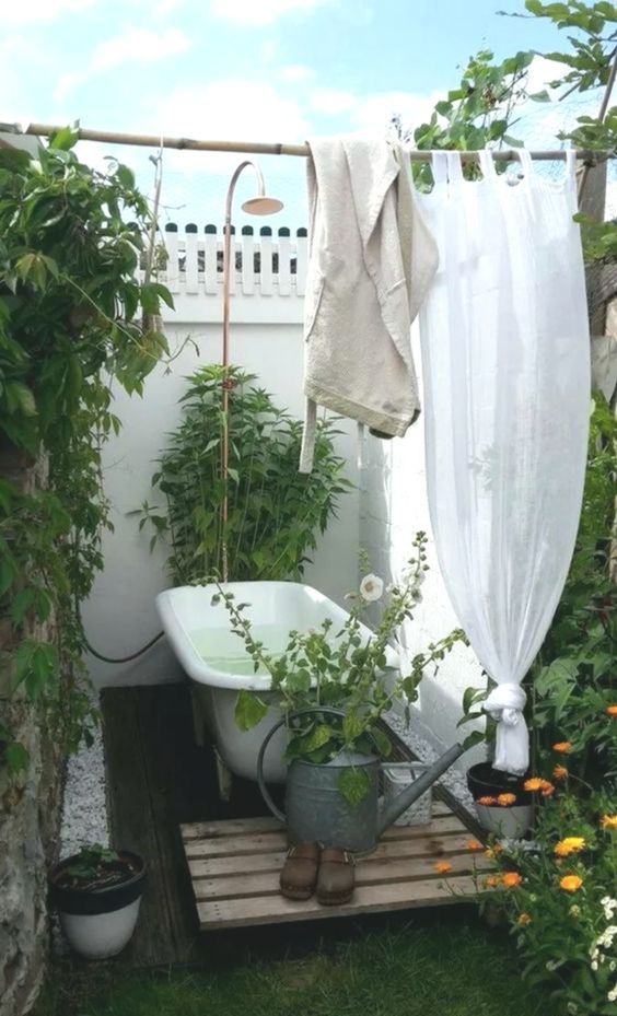 a simple rustic outdoor bathroom with potted greenery and blooms, a vintage tub on a wooden deck and walls and curtains for privacy