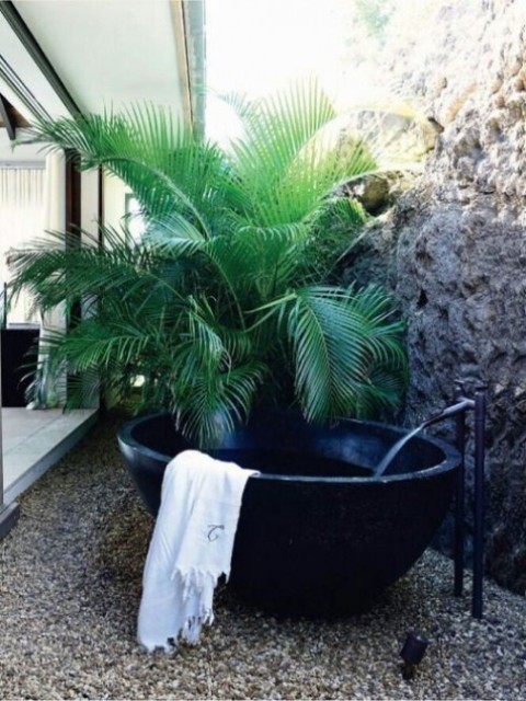 a simple yet gorgeous outdoor oasis with a potted palm, a black stone tub and pebbles on the ground