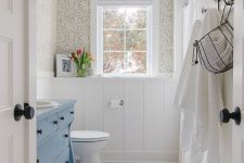 a small vintage bathroom with wallpaper, white penny tiles, a blue vaniyt, white appliances and white textiles