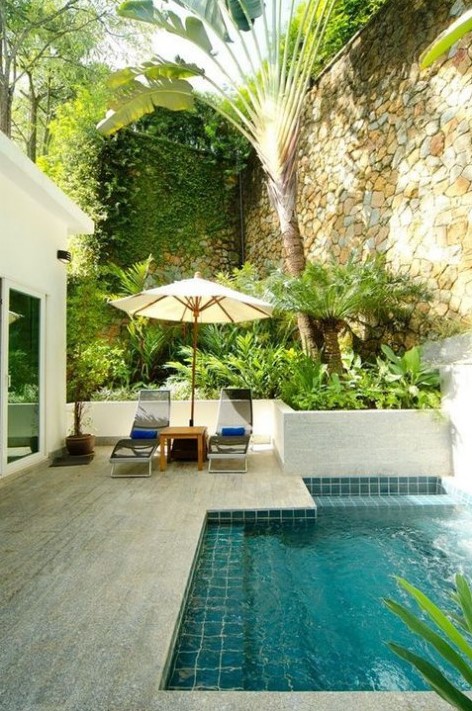 a stone clad pool deck with loungers and an umbrella plus a small backyard pool clad with turquoise tiles inside