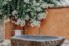 a stunning outdoor bathroom with a carved stone tub placed on sand, rocks and lush blooms over the tub
