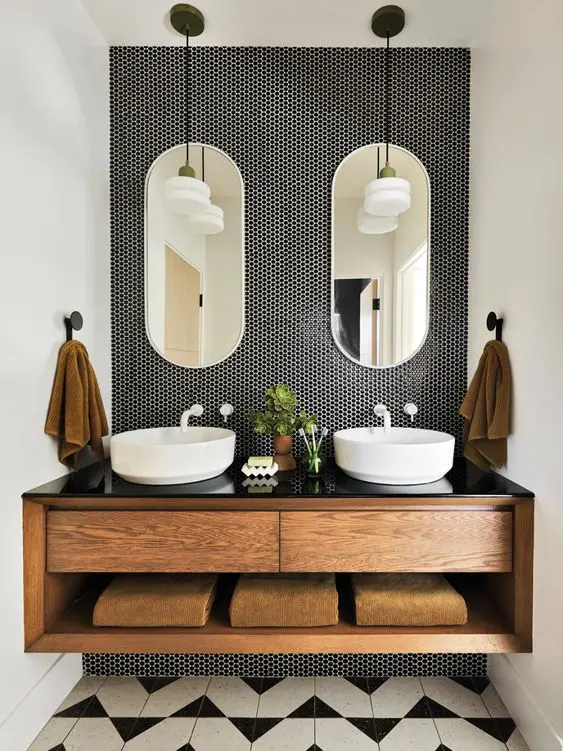 a stylish sink space with black penny tiles, a built-in stained vanity, sinks, oval mirrors, pendant lamps and rust towels