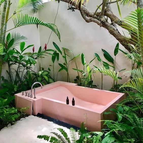 a tropical outdoor bathroom with a comfortable pink tub, lots of greenery growing and white pebbles