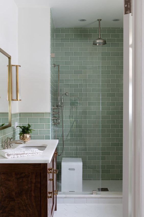 a vintage bathroom with light green subway tiles in the shower, a dark-stained vanity, a mirror and wall lamps