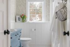 a vintage bathroom with wallpaper, white paneling, white penny tiles on the floor, a blue vanity, some racks