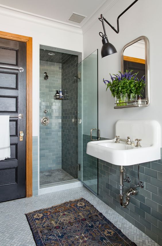 A vintage eclectic bathroom done with grey green subway tiles, a wall mounted sink, a mirror and a wall lamp