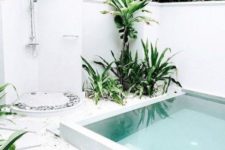 a white tropical backyard with an outdoor shower with pebbles, tropical plants and trees growing and a plunge pool clad in white
