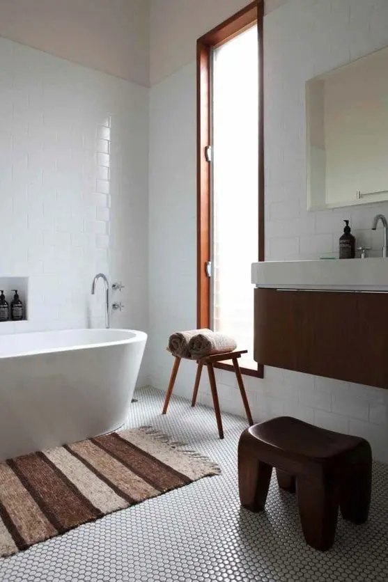 an ethereal mid-century modern bathroom with white penny and subway tiles, an oval tub, a floating vanity and a wooden stool