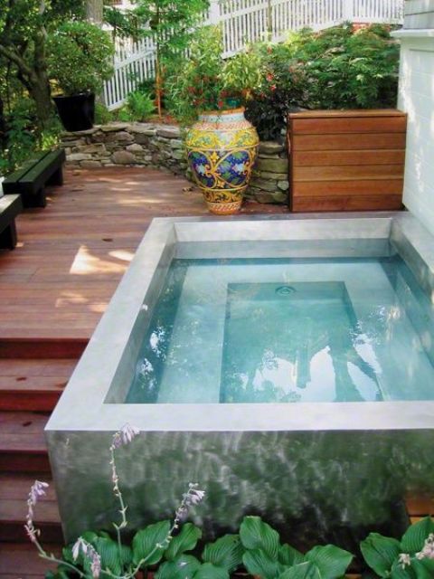 an outdoor space with a wooden deck and a plunge pool, some greenery and blooms around and a couple of benches