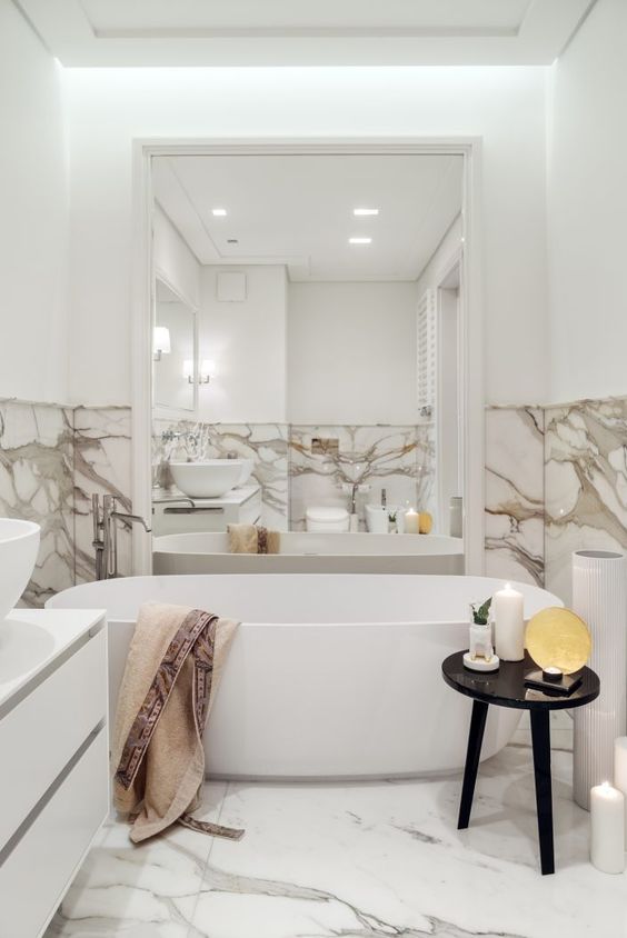 a modern refined bathroom done with white marble, an oval tub and a large mirror in a white frame