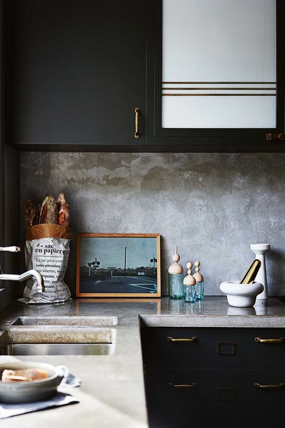 a black kitchen with brass handles, a grey concrete countertop and a matching backsplash for a moody touch