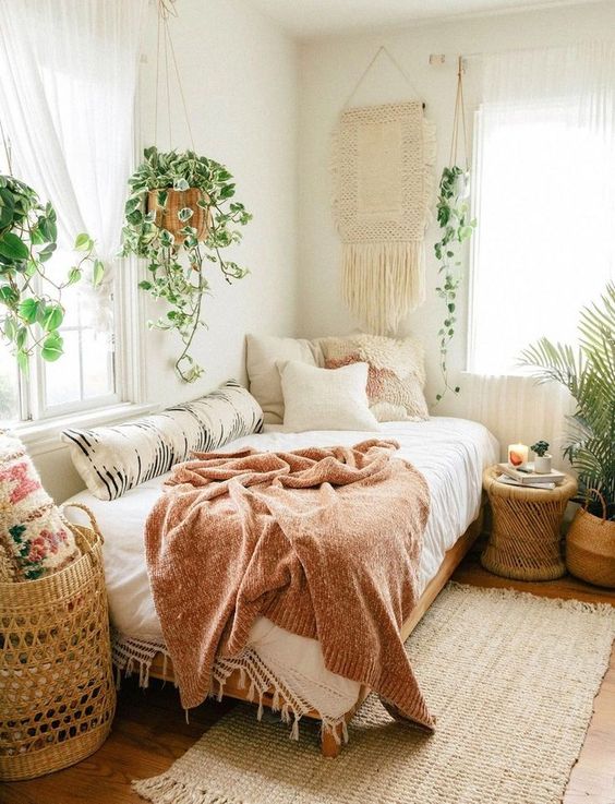 a boho bedroom with a macrame hanging, lots of potted plants, a basket, a wicker table and pretty pillows
