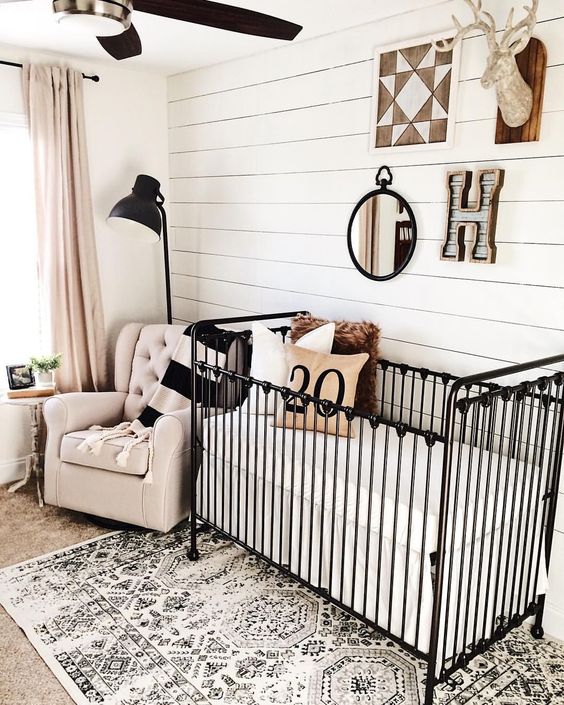 a farmhouse nursery with a white plank wall, a printed rug, a black metal crib and some more black touches for drama