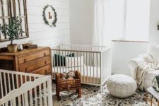 a neutral farmhouse nursery with a printed rug, white beds, a white leather ottoman, stained wooden furniture and a beaded chandelier