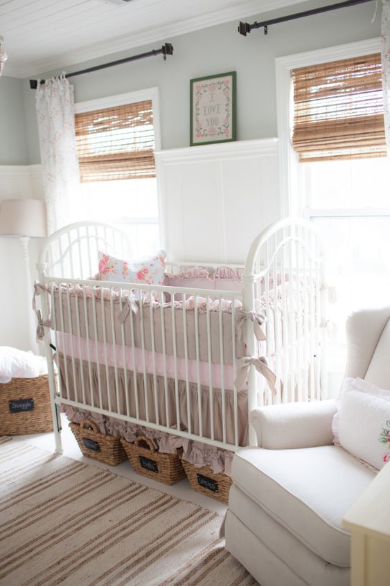 a vintage farmhouse nursery with white walls, woven shades, baskets, a white vintage crib with ruffles and bows