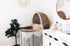 a wild boho nursery with a wicker bassinet and a macrame garland, a white dresser with wooden inserts and leather pulls, a macrame gallery wall and potted greener