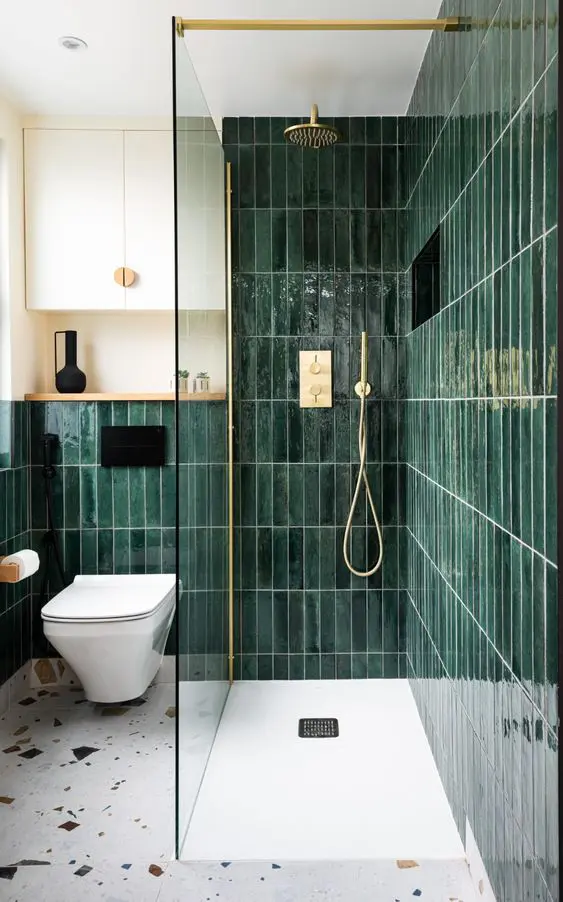 a beautiful bathroom with dark green skinny tiles, a terrazzo floor, a toilet and some gold and brass appliances