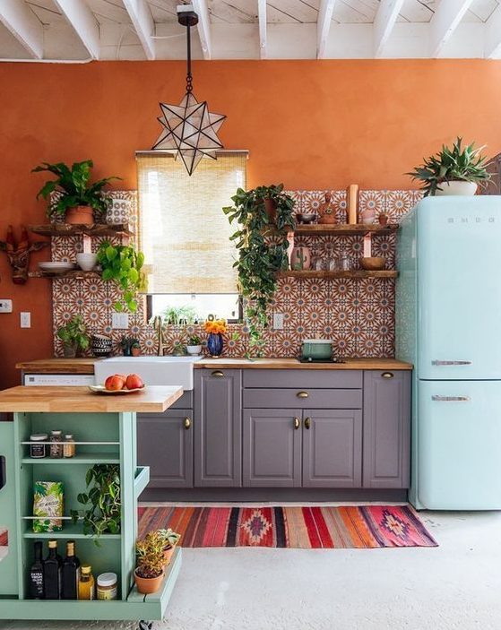 A bright maximalist kitchen with grey cabinets, an aqua colored kitchen island and a matching fridge, rust walls and bright tiles