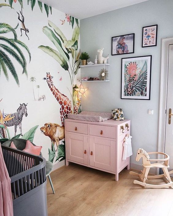 a bright tropical nursery with a pink changing table, a bright jungle print wall, bright artworks and a grey crib with a pink canopy