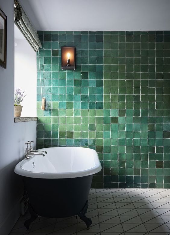a catchy bathroom with a very special green tile wall in various shades, a vintage black tub and a mosaic floor