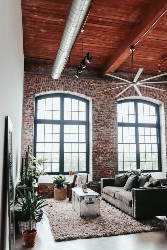 a chic industrial living room with brick walls, a wooden ceiling, exposed pipes, a metal chest, lots of greenery