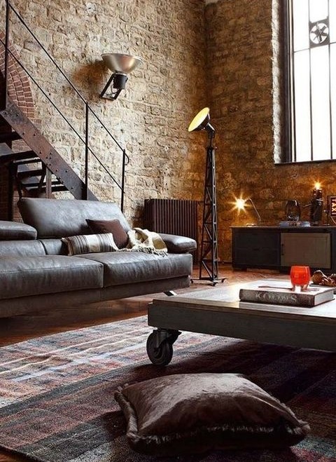 a chic industrial living room with stone walls, a leather sofa, a wood and metal table, a metal staircase and lamps here and there
