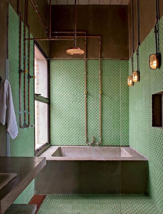 a colorful industrial bathroom with green polka dot tile walls, a concrete bathtub, exposed piping and vintage lamps